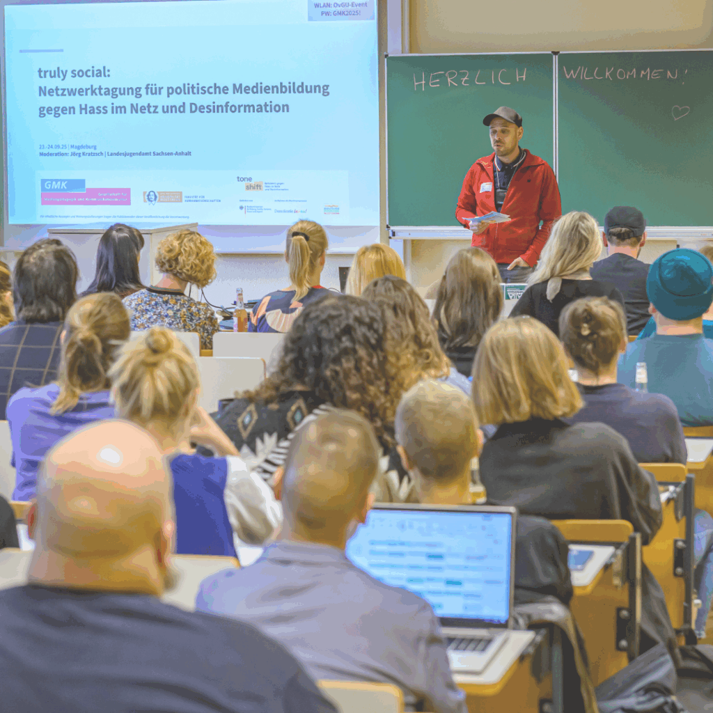 Eine Person steht in einem Seminarraum vor einer Gruppe von Teilnehmenden und spricht. Auf einer Leinwand ist der Titel „truly social: Netzwerktagung für politische Medienbildung gegen Hass im Netz und Desinformation“ zu sehen. An der Tafel daneben steht in Kreideschrift „Herzlich willkommen!“.