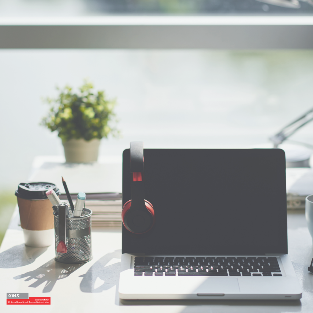 Moderner Arbeitsplatz mit aufgeklapptem Laptop, roten Kopfhörern am Bildschirm, Kaffeebecher und Stiftehalter auf einem hellen Schreibtisch, im Hintergrund eine Pflanze bei Tageslicht.