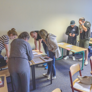 Abgebildet ist ein Workshopsetting in einem Seminarraum: Mehrere Menschen, aufgeteilt in zwei Gruppen, beugen sich gemeinsam über Tische auf denen Arbeitsmaterialien verteilt sind. Sie sehen vertieft in die Materialien und konzentriert aus.