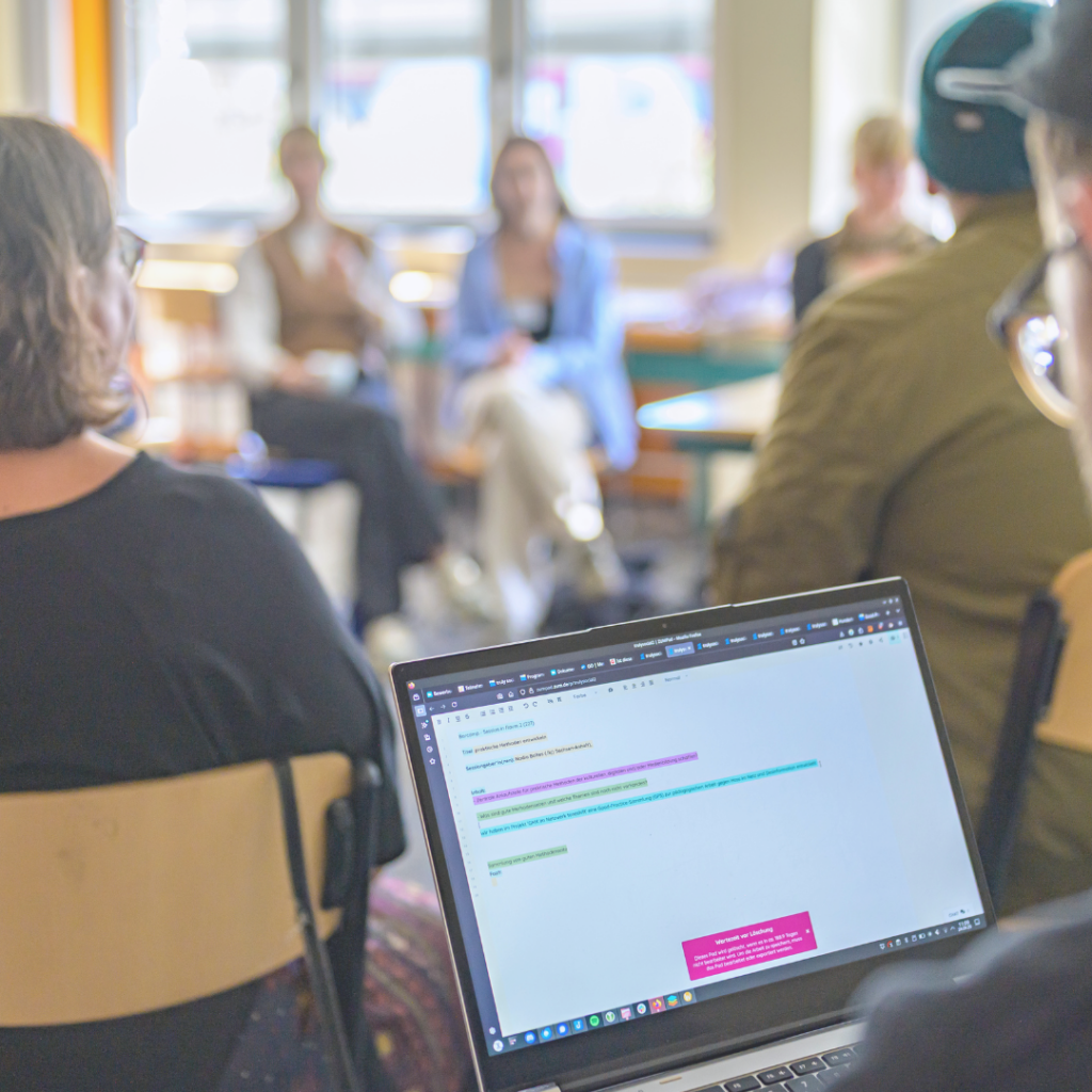Unscharfe Aufnahme einer Gruppendiskussion in einem hellen Raum. Im Vordergrund ist ein aufgeklappter Laptop zu sehen, auf dessen Bildschirm farbig markierte Notizen geöffnet sind. Im Hintergrund sitzen mehrere Personen im Kreis auf Stühlen und hören zu oder sprechen miteinander.